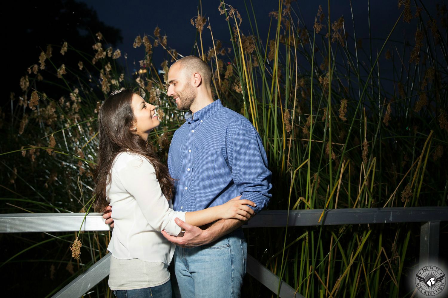 Brown haired Latino girl wearing white elbow length sweater and blue jeans happily looks into the eyes of a tall guy wearing a button up blue dress shirt and blue jeans in front of a railing on a boat dock with tall green reeds in the background at dusk on Ladybird Lake in this friendly engagement photo in downtown Austin, Texas.
